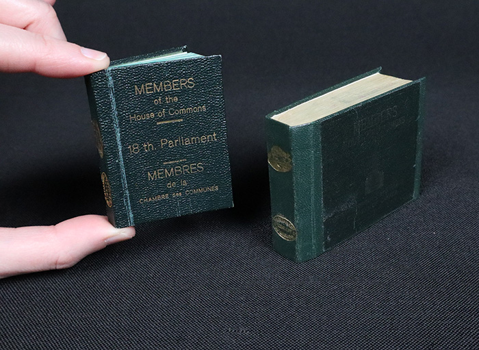 Two small books, bound in dark green, standing upright. The smallest volume is being held between someone’s thumb and forefinger to give an idea of scale.